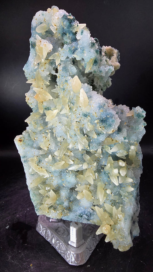 A mineral specimen featuring pale blue-green and cream-colored crystals on a dark background, with a small carved gray stone piece visible at the lower right.